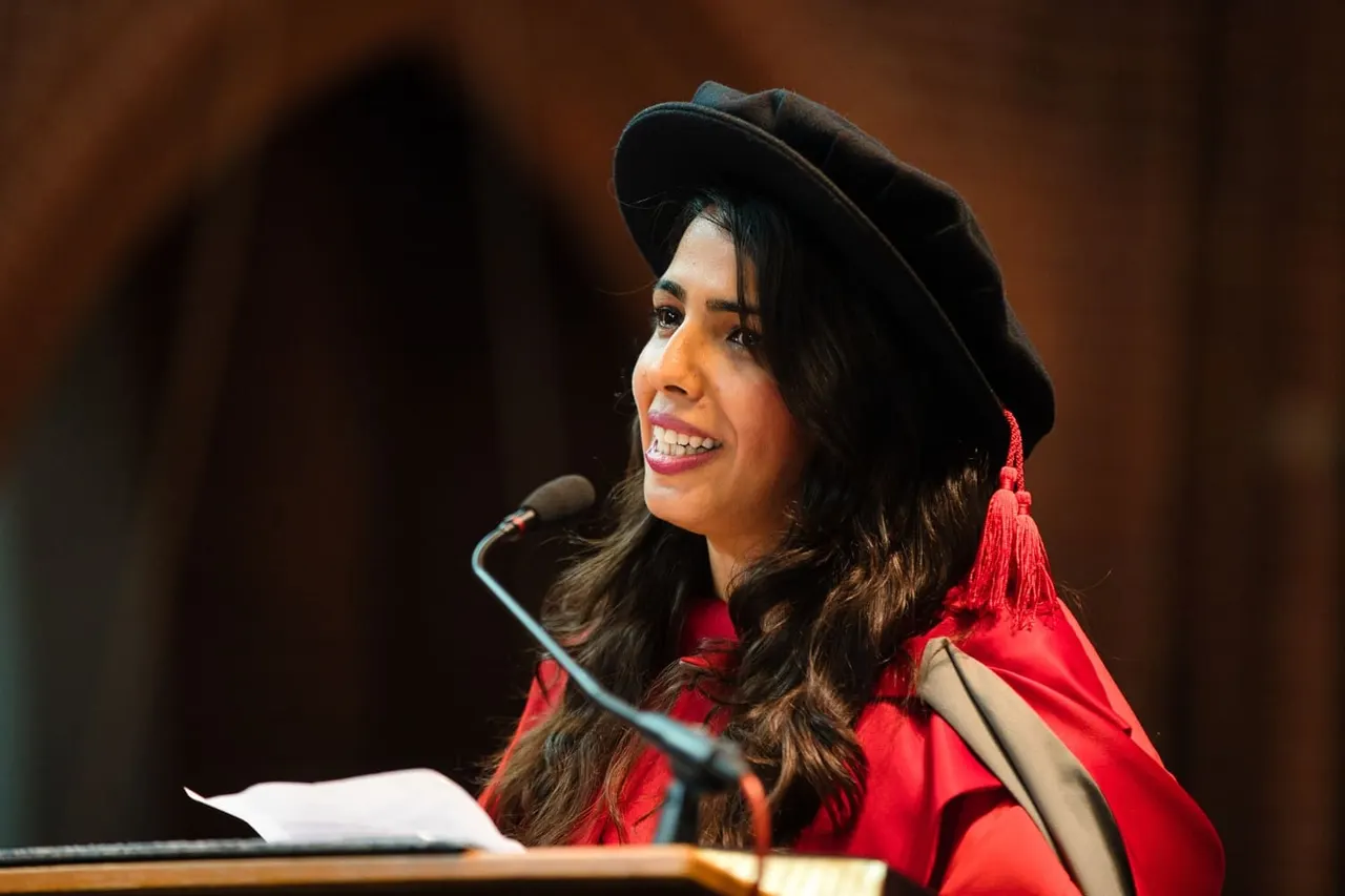 Woman in academic regalia speaking at a microphone during a graduation ceremony.