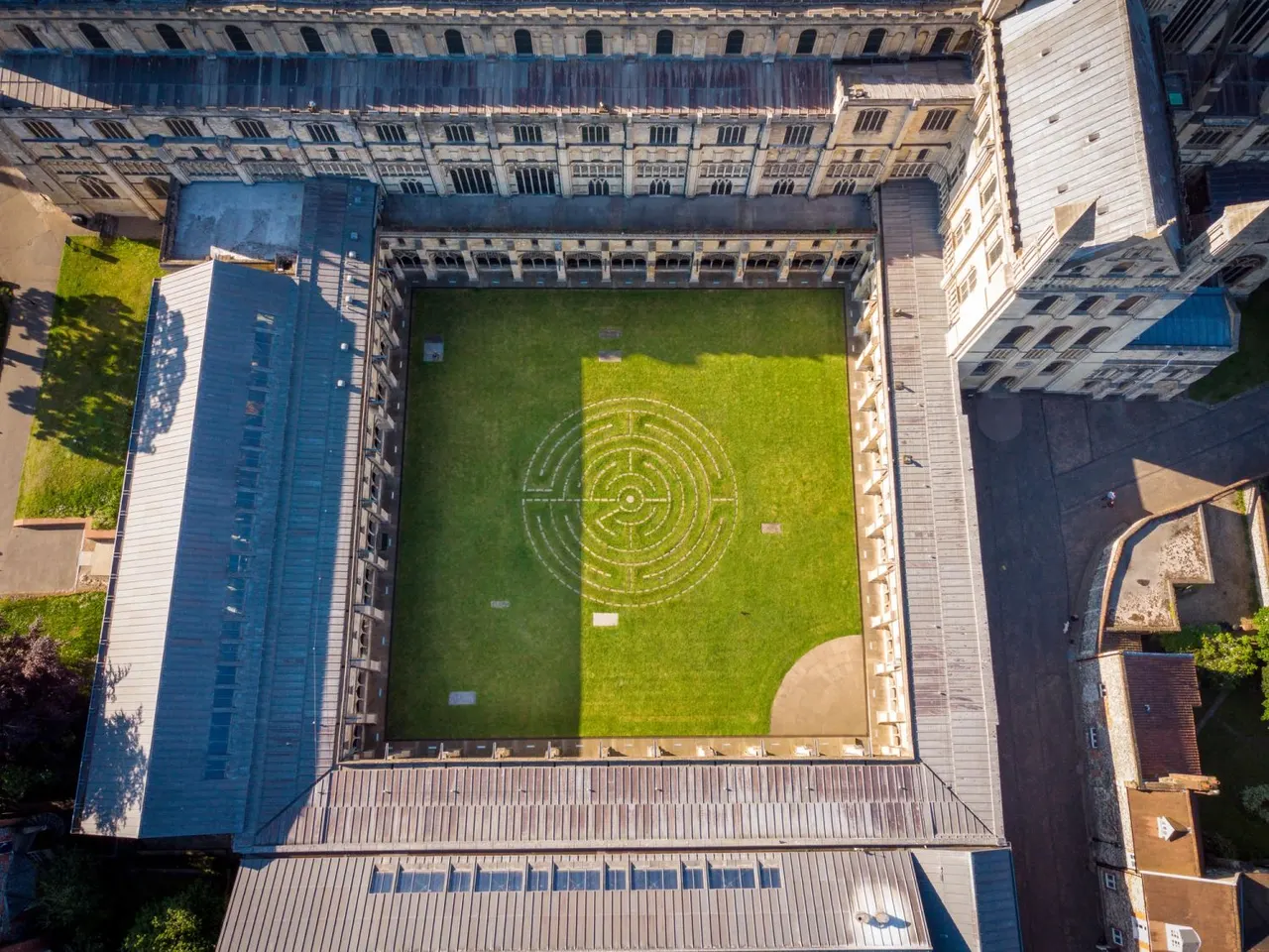Aerial view of a courtyard garden with circular maze pattern on grass, surrounded by historic stone buildings.