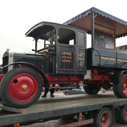 Vintage steam-era commercial vehicle with red wheels and black cabin on a transport trailer.