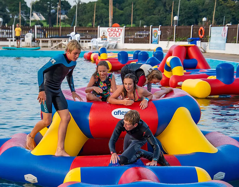 Children and young people playing on colourful inflatable obstacle course equipment in an outdoor swimming pool.