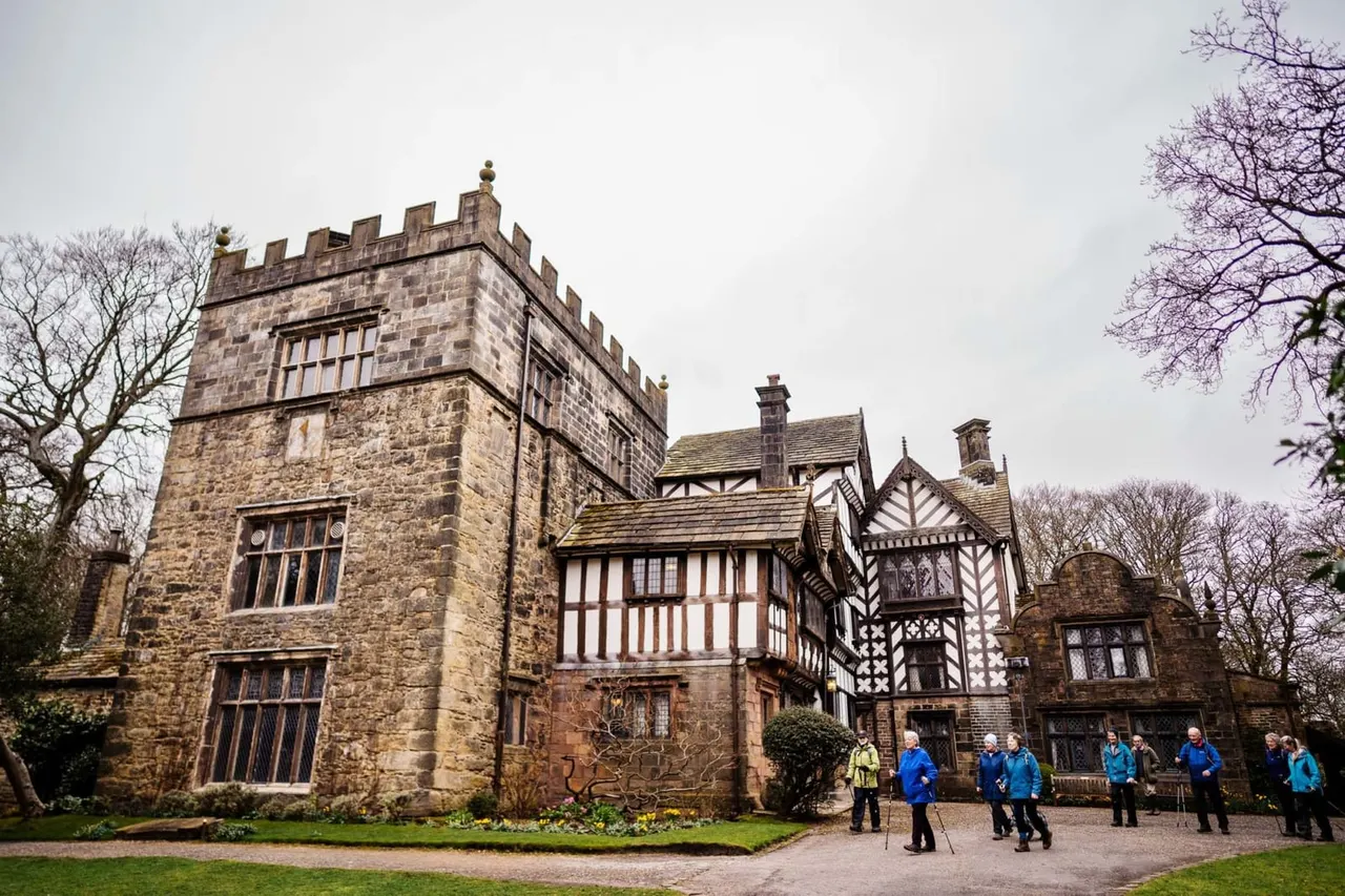 A group of visitors in colourful outdoor clothing stand in the grounds before a historic stone tower and half-timbered manor house.