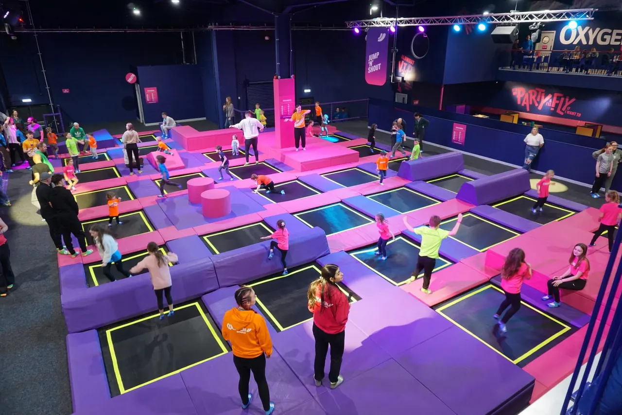 Children and adults bouncing on a large indoor trampoline park with neon pink and purple padded sections under bright stage lighting.