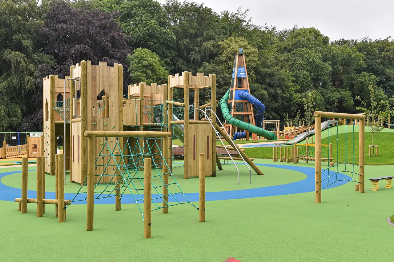 Large wooden castle-themed play structure with slides, climbing nets and rope features on bright green safety surfacing, surrounded by trees.