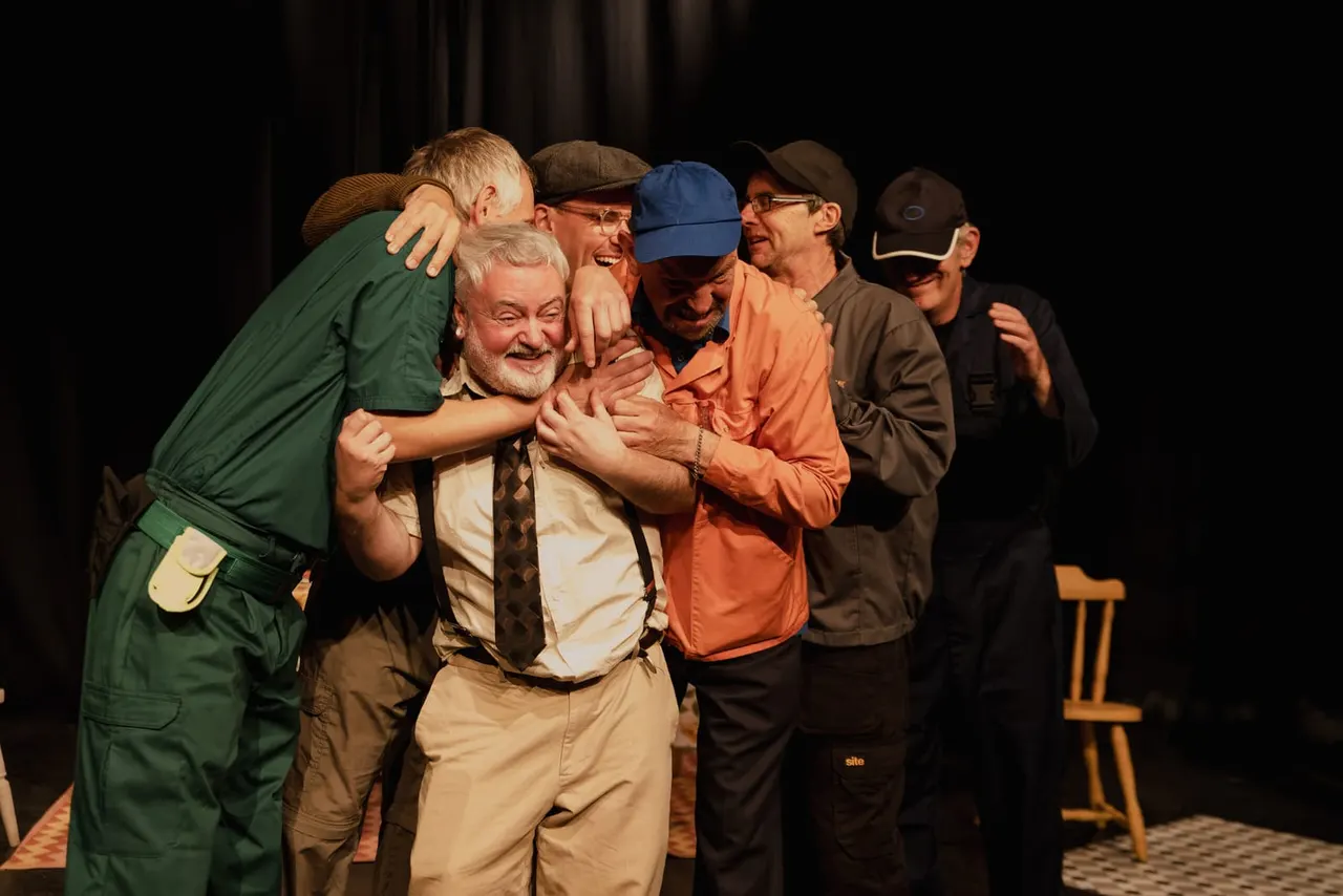 Group of men in outdoor clothing embracing and laughing together on stage during a theatrical performance.
