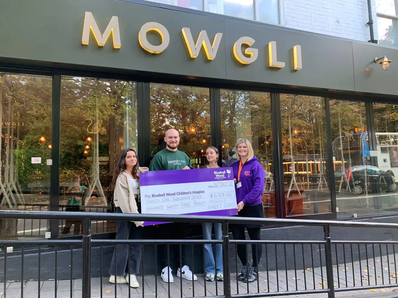 Four people holding a large purple charity cheque outside a restaurant storefront.