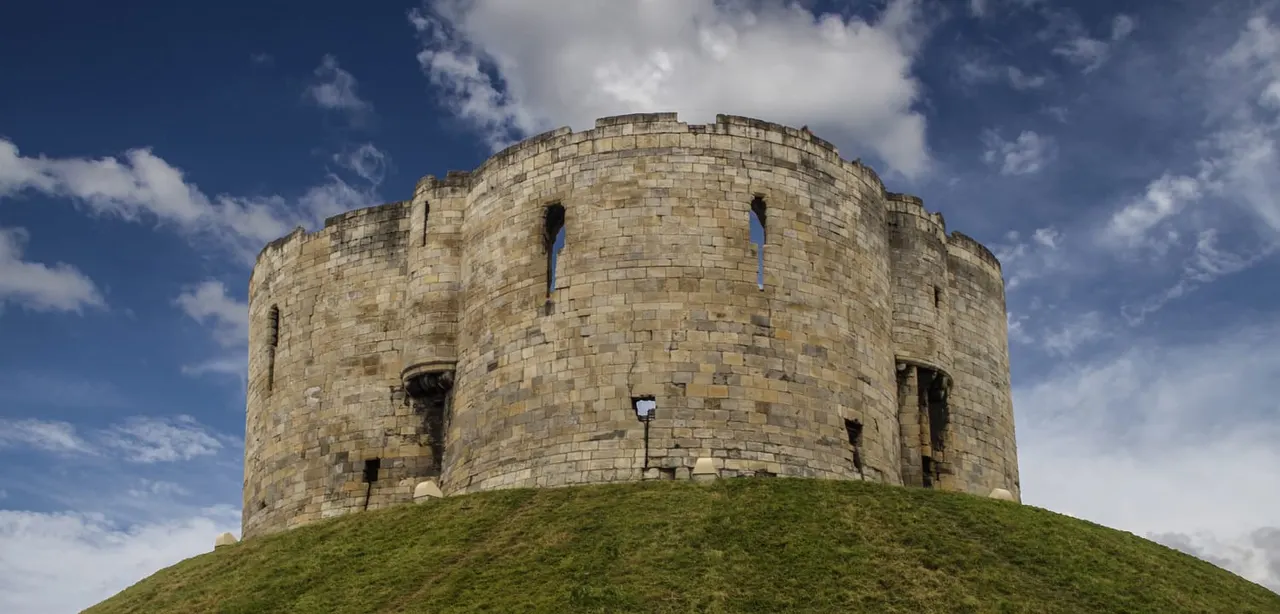 Clifford's Tower, a circular stone medieval fortress, sits on a grass mound under blue sky with white clouds.