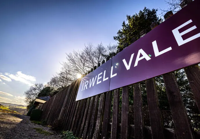 A purple and white sign reading "Irwell Vale" mounted on a dark wooden fence, with trees and countryside visible beyond.