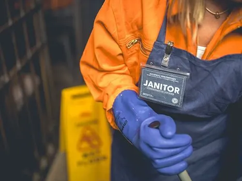 Janitor wearing orange and blue uniform holding cleaning supplies with ID badge visible.