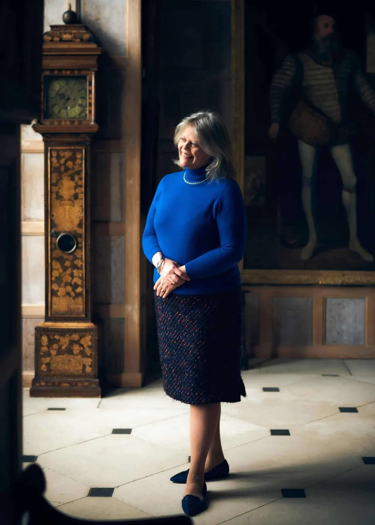 A woman in a bright blue jumper stands in an ornate interior hall beside an inlaid wooden longcase clock, with other visitors visible in the background.