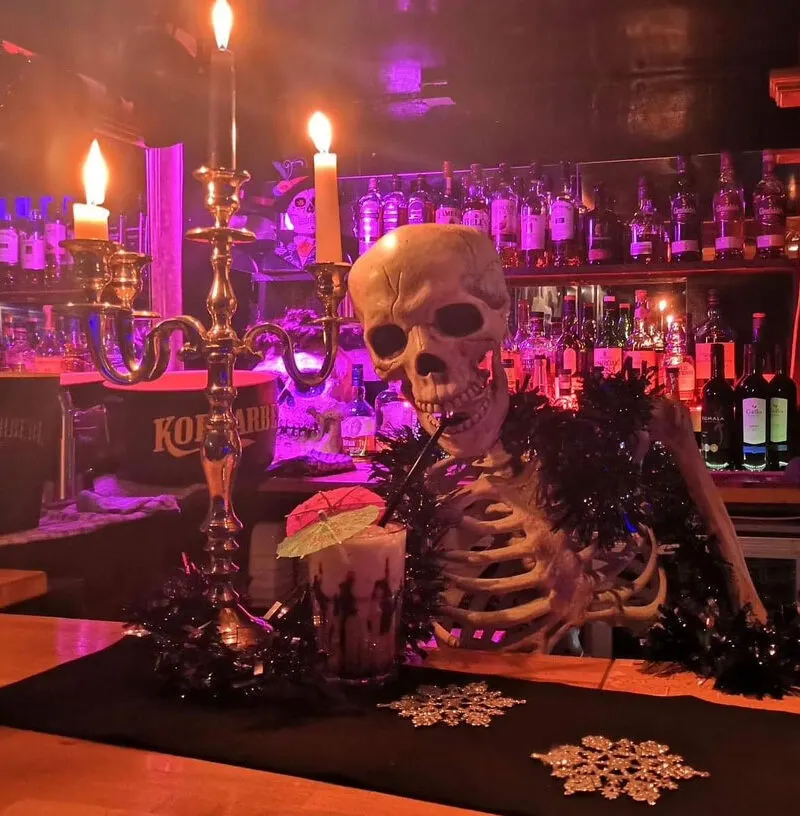 A skeleton decoration with lit candles and cocktail glass at a bar counter, surrounded by purple and orange lighting with spirit bottles on shelves behind.