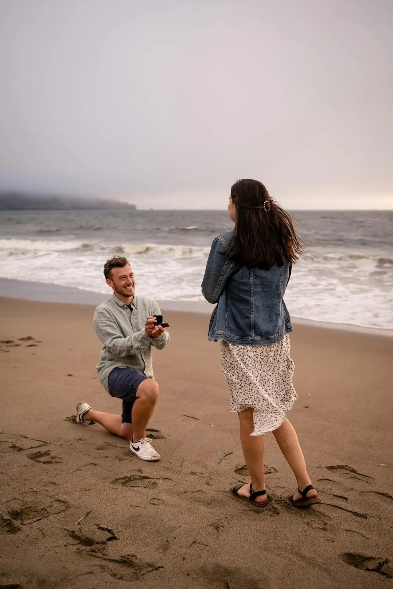 Man on one knee proposing to a woman on a sandy beach with waves and cliffs behind them.
