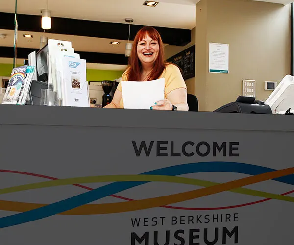 A woman with red hair wearing a yellow top smiles behind a reception desk marked "Welcome" at West Berkshire Museum.