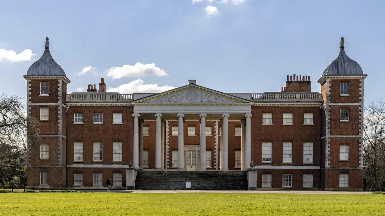 Red brick Georgian mansion with domed towers, columned portico and manicured lawn under blue sky with white clouds.