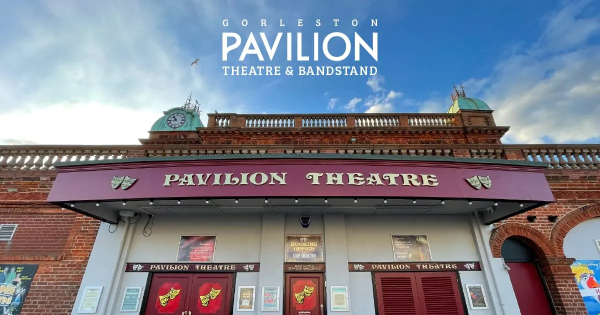 Victorian brick building with clock towers and green domes, purple theatre marquee, and red entrance doors under blue sky.