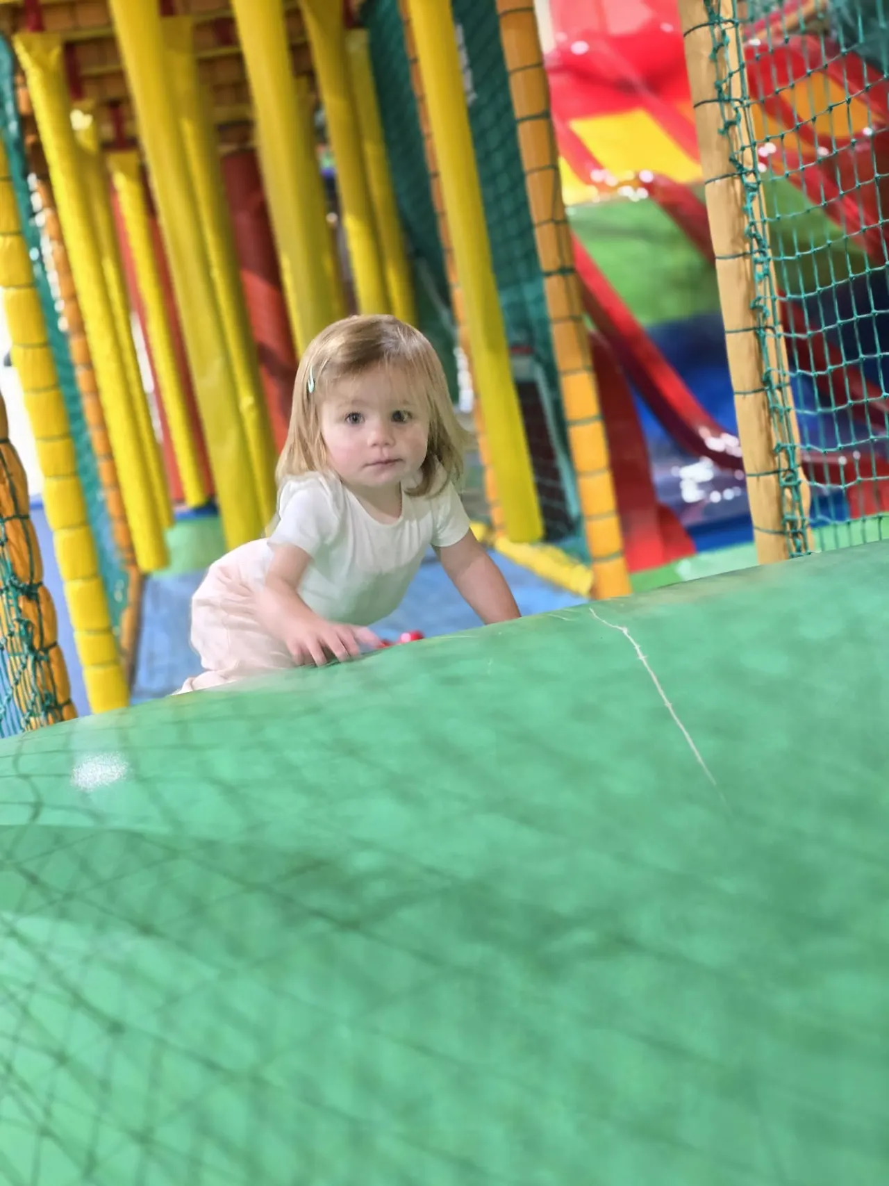 Toddler crawling through a green soft play tunnel surrounded by colourful inflatable structures.