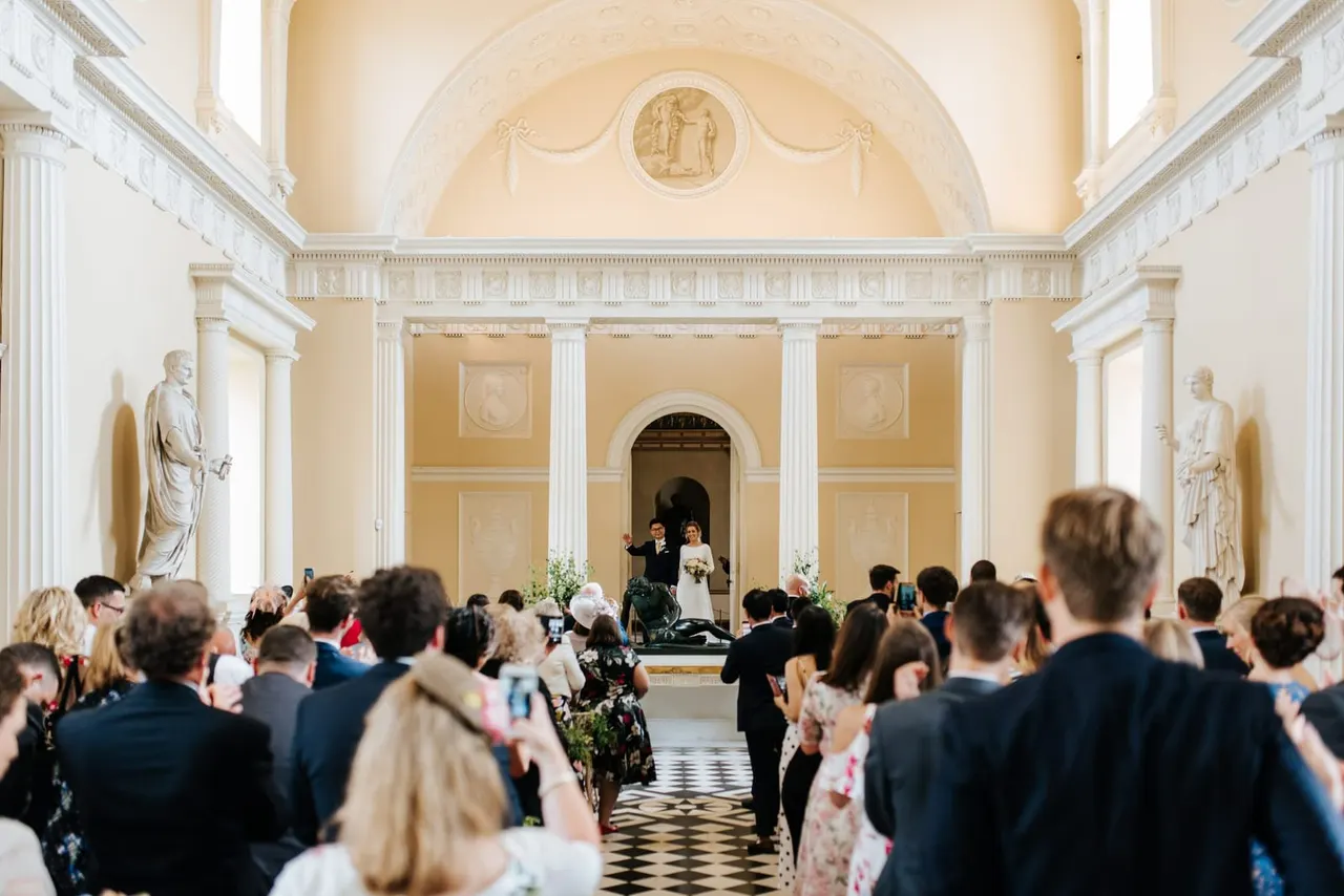 Wedding ceremony in a grand neoclassical hall with cream and gold walls, white columns, and classical sculptures, bride and groom at altar whilst guests watch.