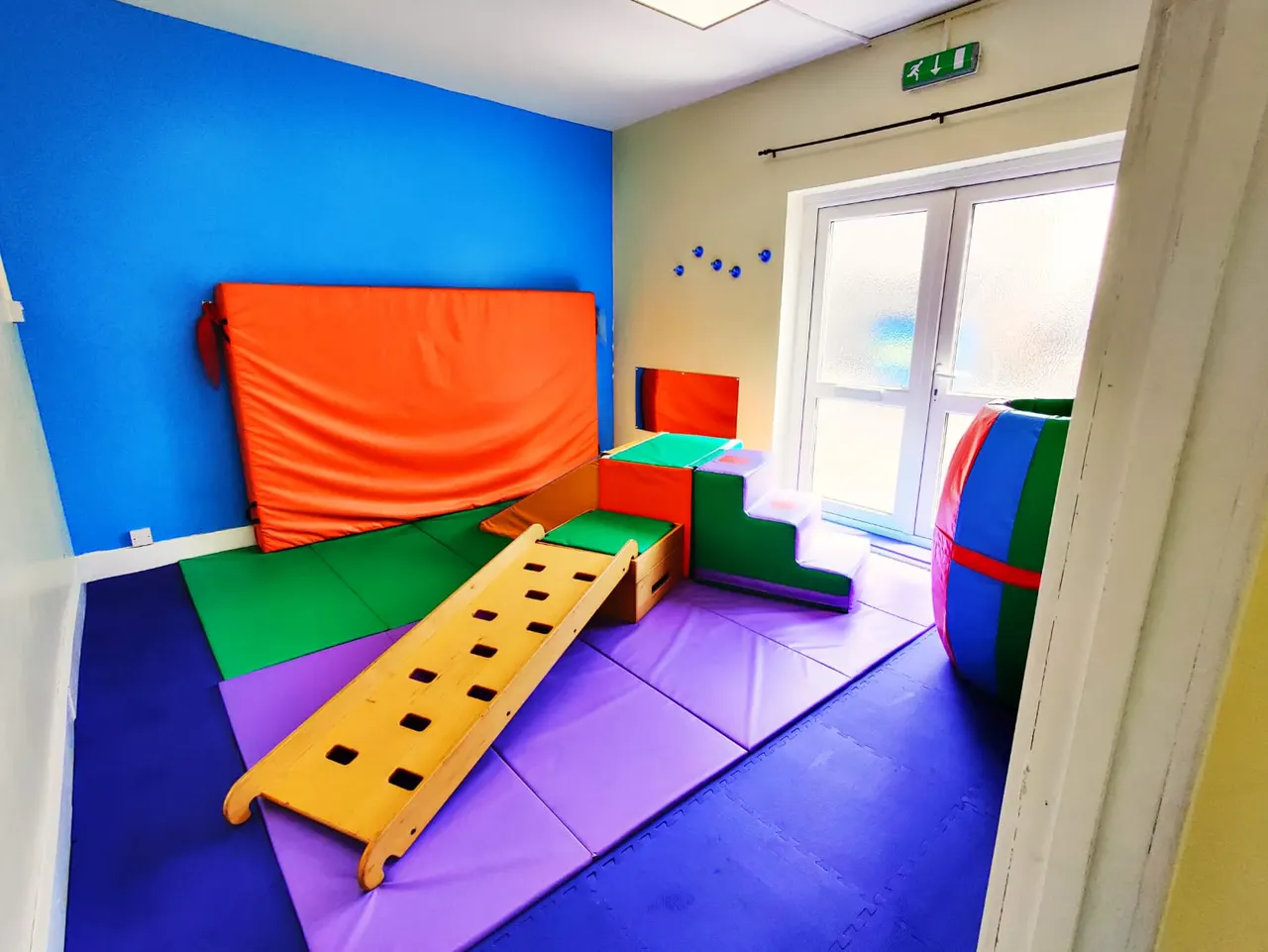 Colourful indoor soft play room with padded equipment including foam blocks, climbing structures and a yellow balance beam on purple matting.