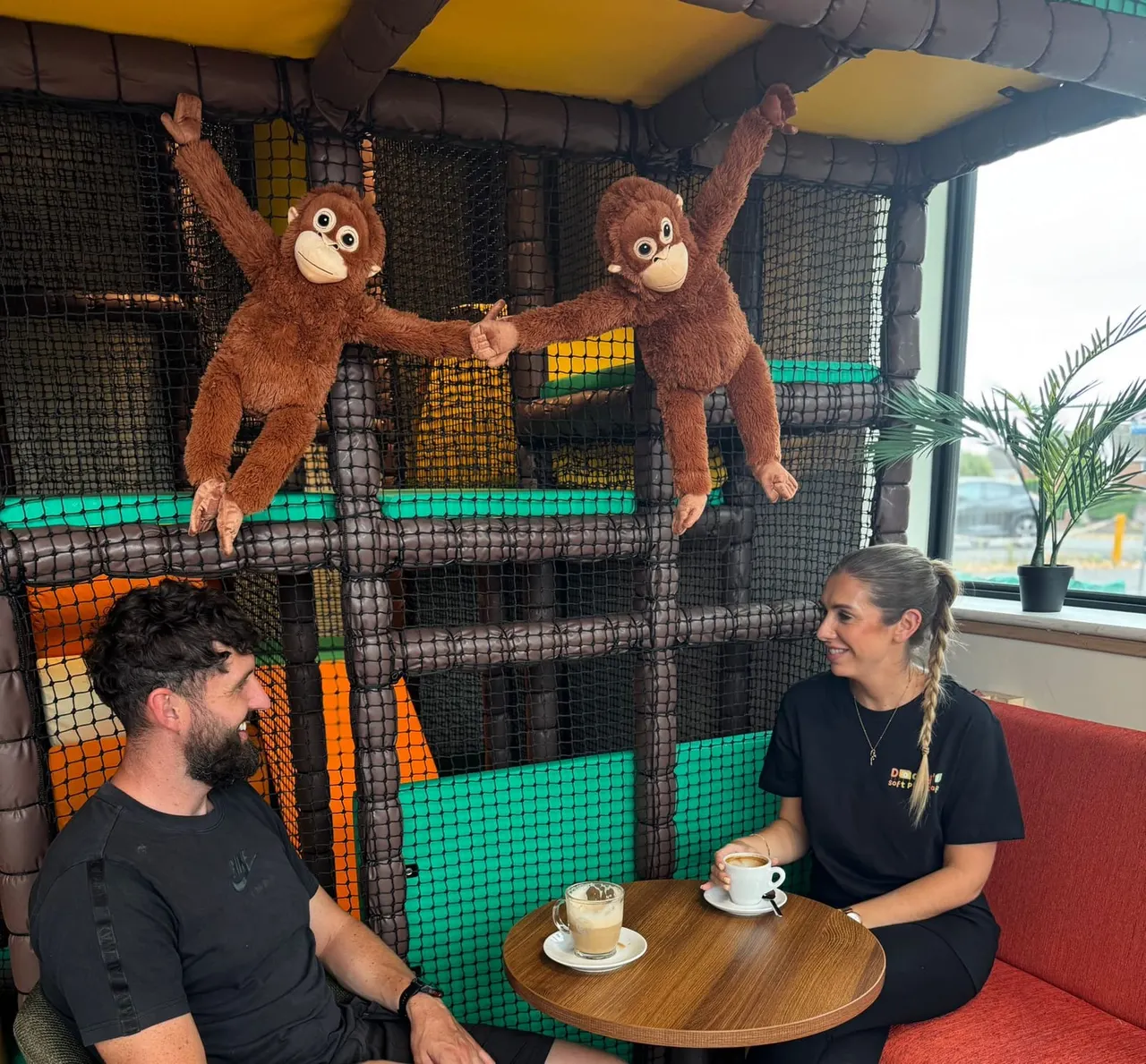 Two adults sit at a table with drinks while oversized primate costume characters perform above them inside an indoor soft play facility.