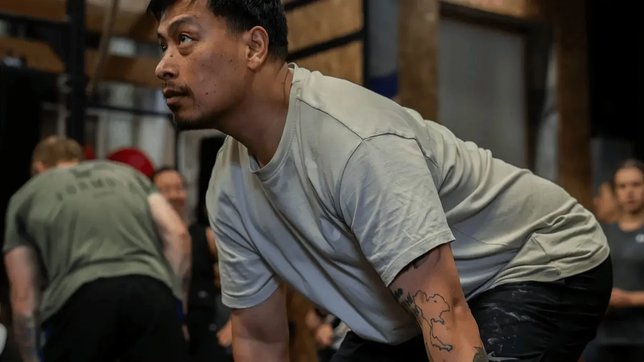 Man in grey t-shirt performing a weightlifting exercise indoors with other gym members watching in the background.