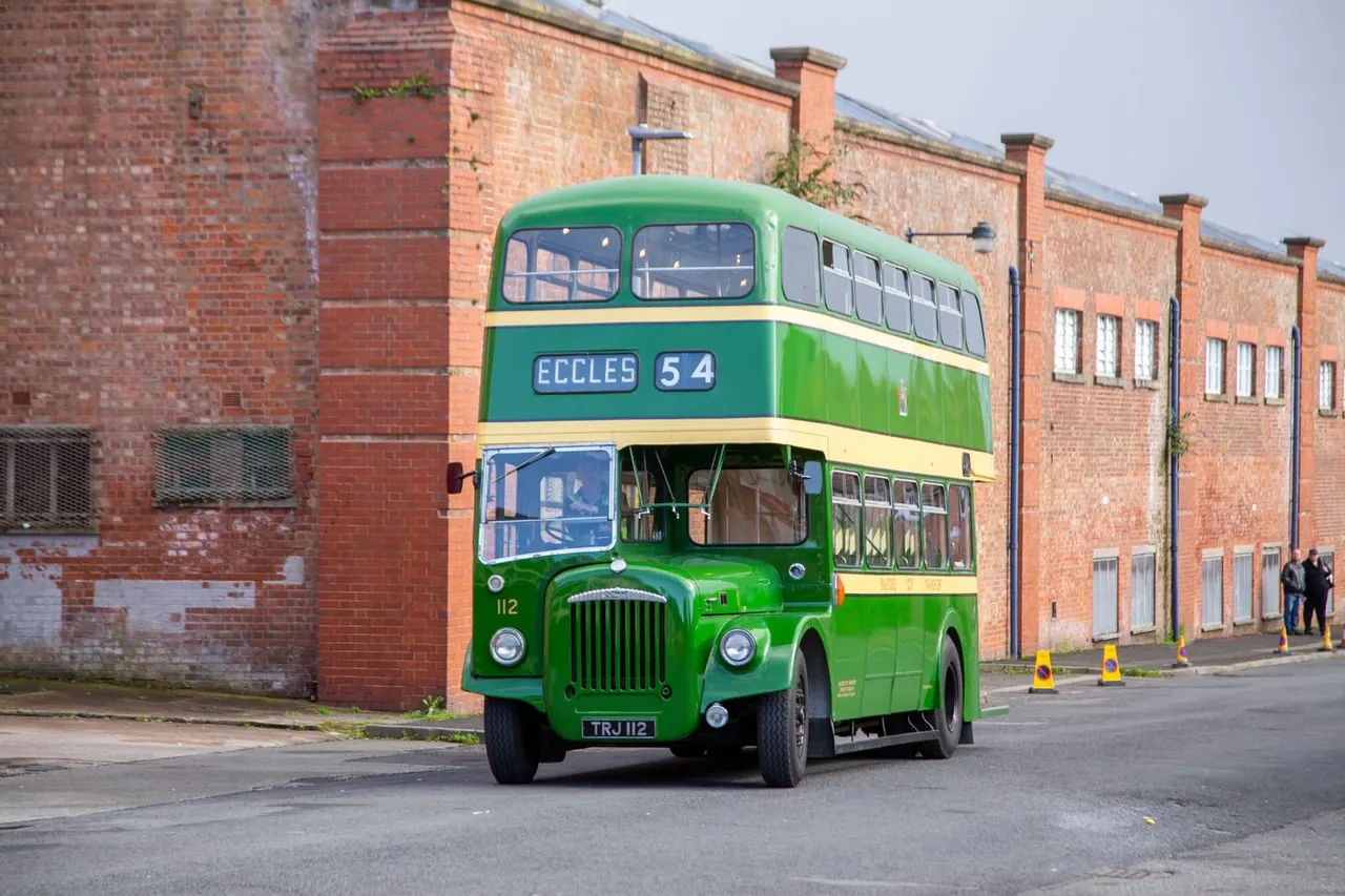Green and cream vintage double-decker bus parked in front of red-brick industrial buildings.