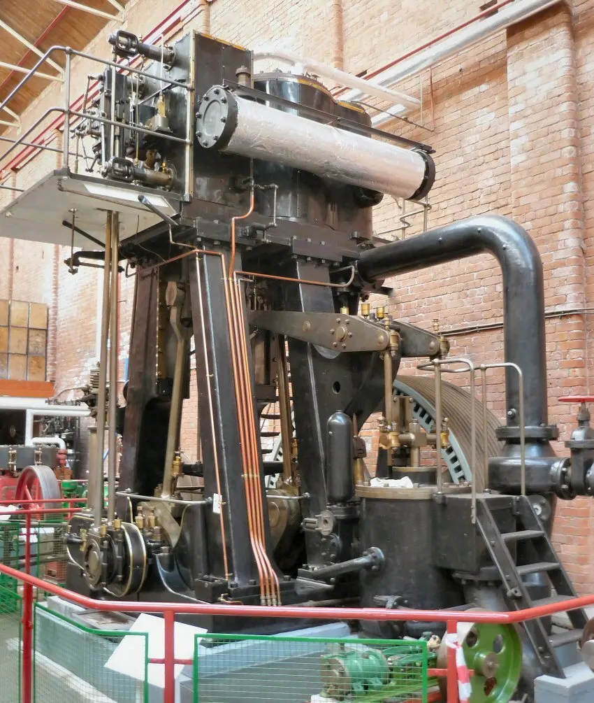 Historic stationary steam engine with cylindrical boiler and mechanical components displayed indoors against a brick wall.