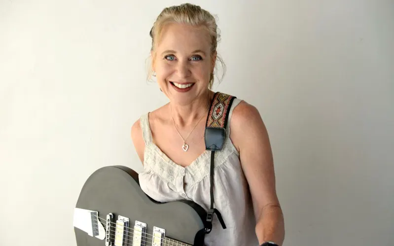 Woman holding an electric guitar against a plain background, smiling at camera.