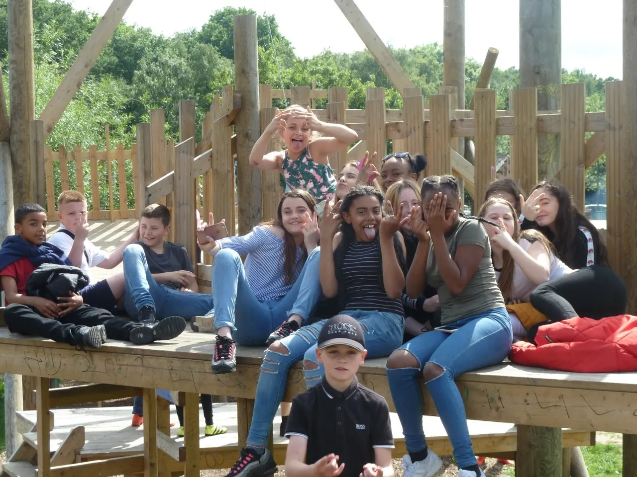 Group of children and young teenagers sitting and posing together on wooden play structure platform with trees in background.