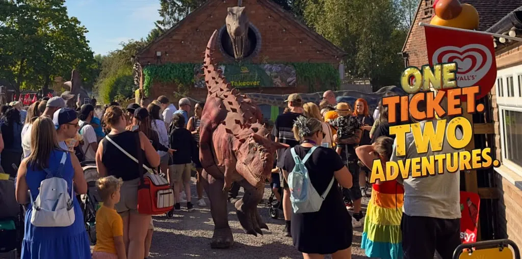 Crowds of visitors gather around a large dinosaur statue at an outdoor leisure venue on a sunny day.