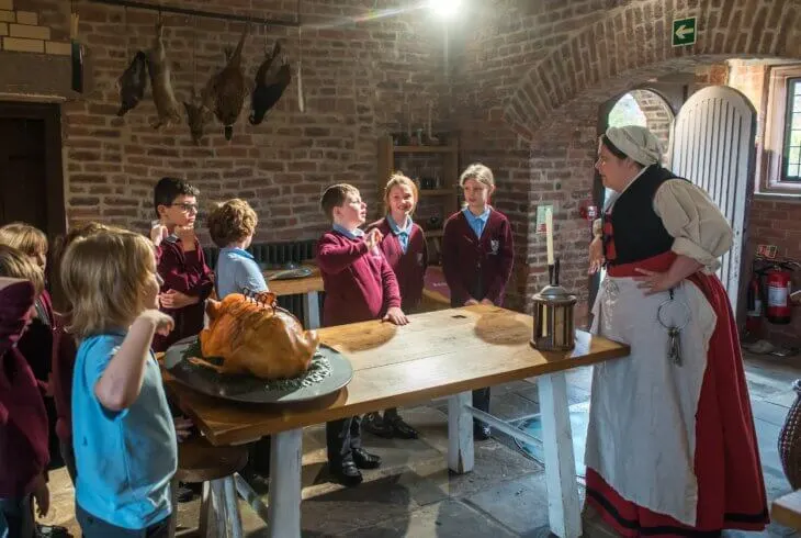 A woman in historical costume explains food preparation to a group of schoolchildren around a wooden table in a brick-built historic kitchen.