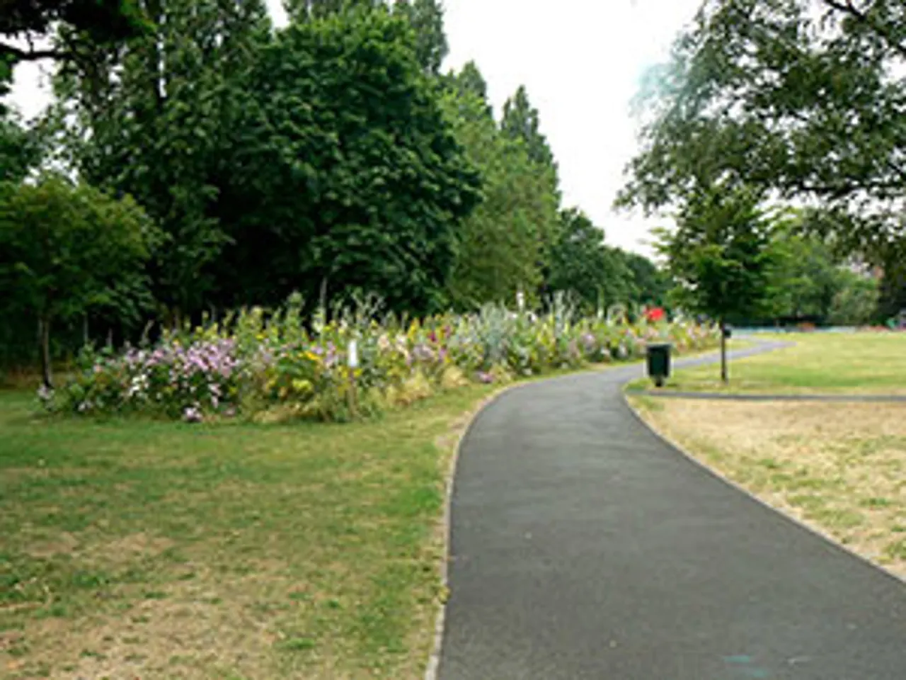 Curved tarmac path through park with flowering borders, mature trees and grass verges on either side.
