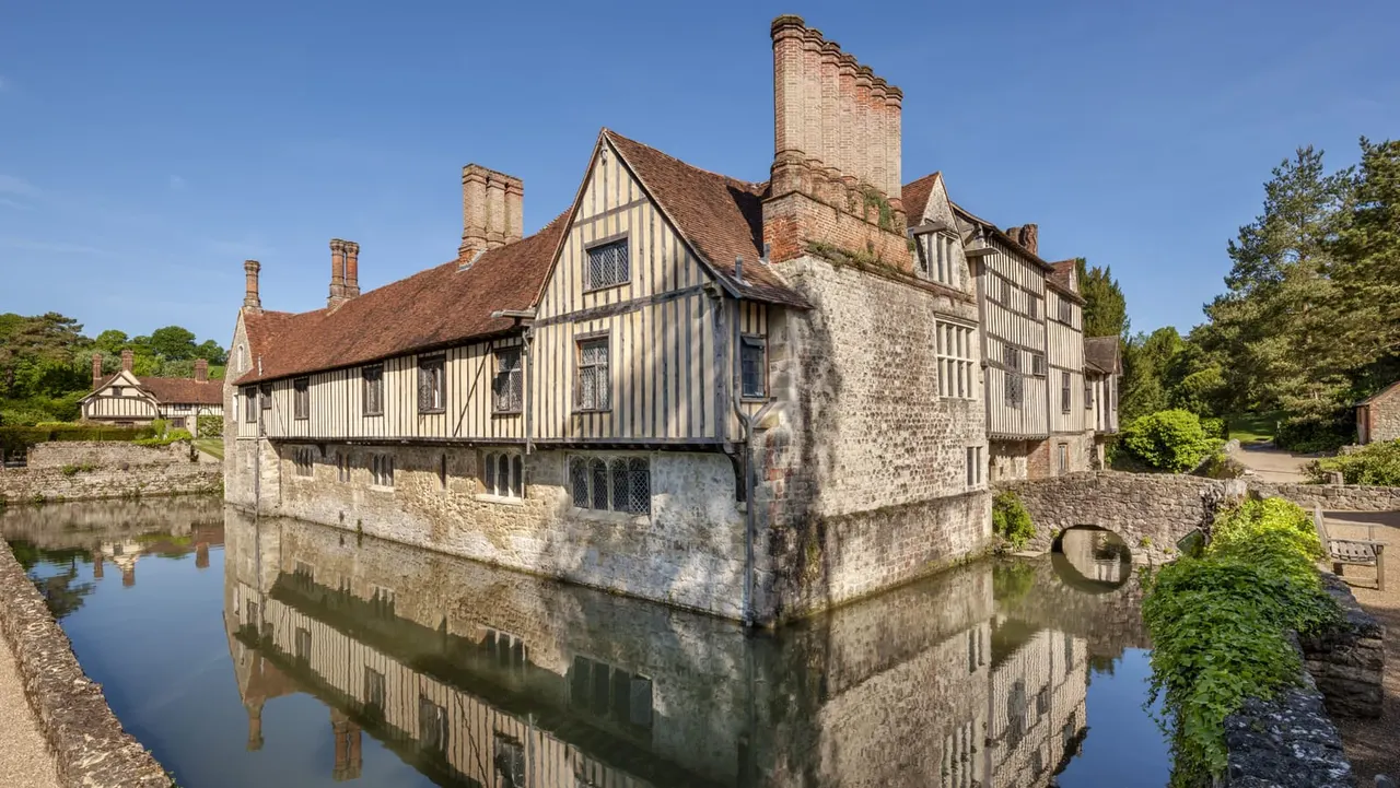 Medieval half-timbered manor house with stone base surrounded by a moat, set within wooded grounds under clear blue sky.
