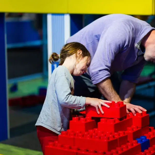 Adult and child building with large red plastic blocks in an indoor play area.