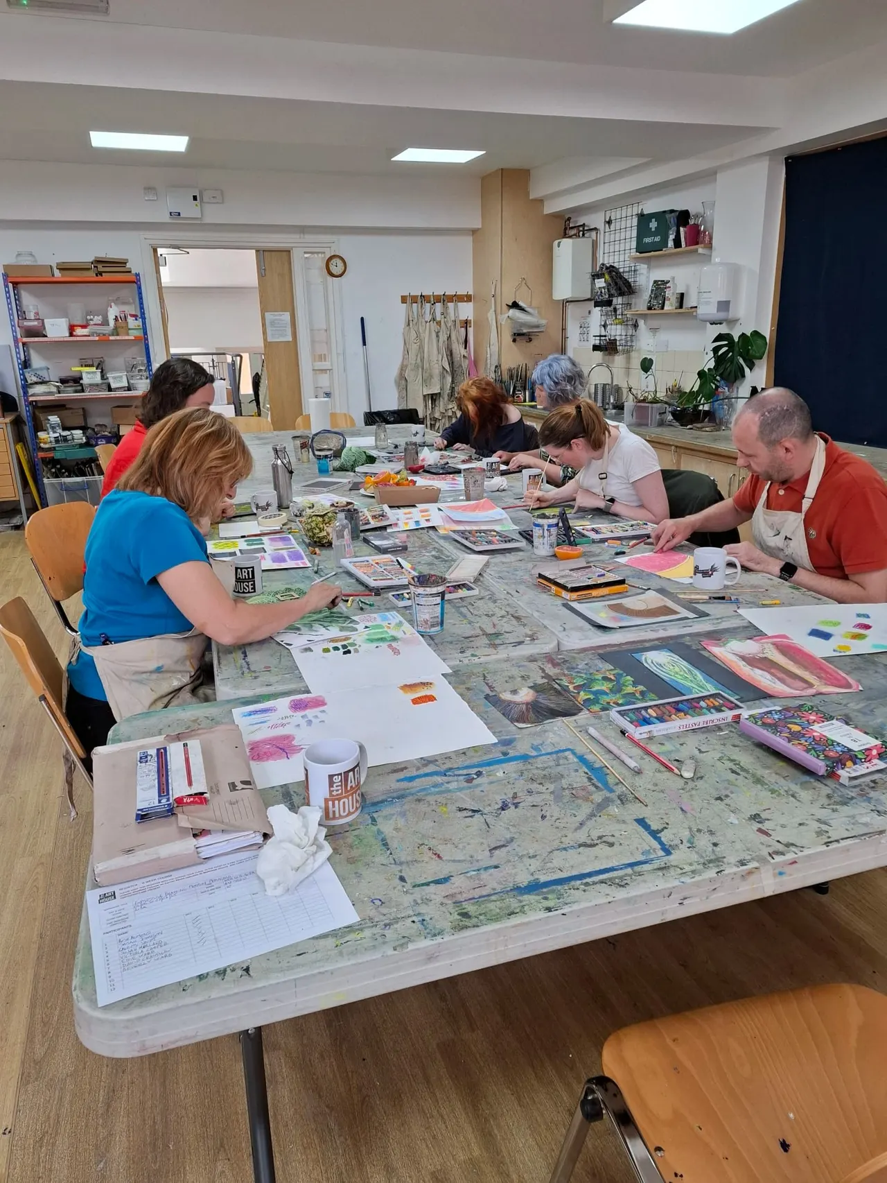A group of five adults working on art and craft projects together around a large table covered with paints, brushes, and papers in a bright studio space.