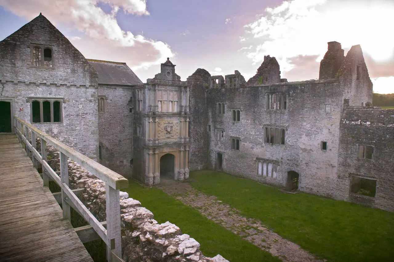 Ruined stone castle with grassy courtyard, surrounded by partially intact walls and towers, viewed from wooden bridge under cloudy sky.