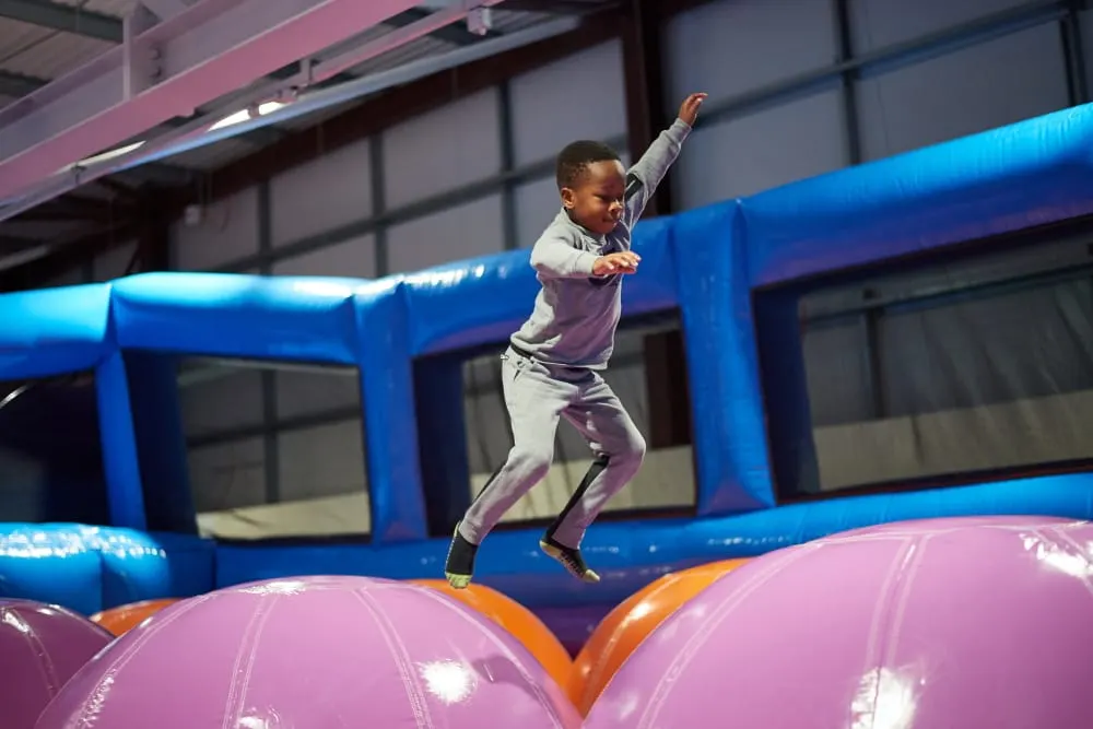 Child bouncing on purple and pink inflatable domes inside an indoor trampoline park with blue structures.