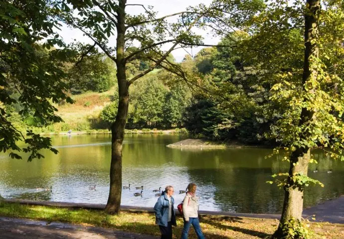 Two visitors walk by a tree-lined river bank with ducks swimming on the water and rolling green hills beyond.