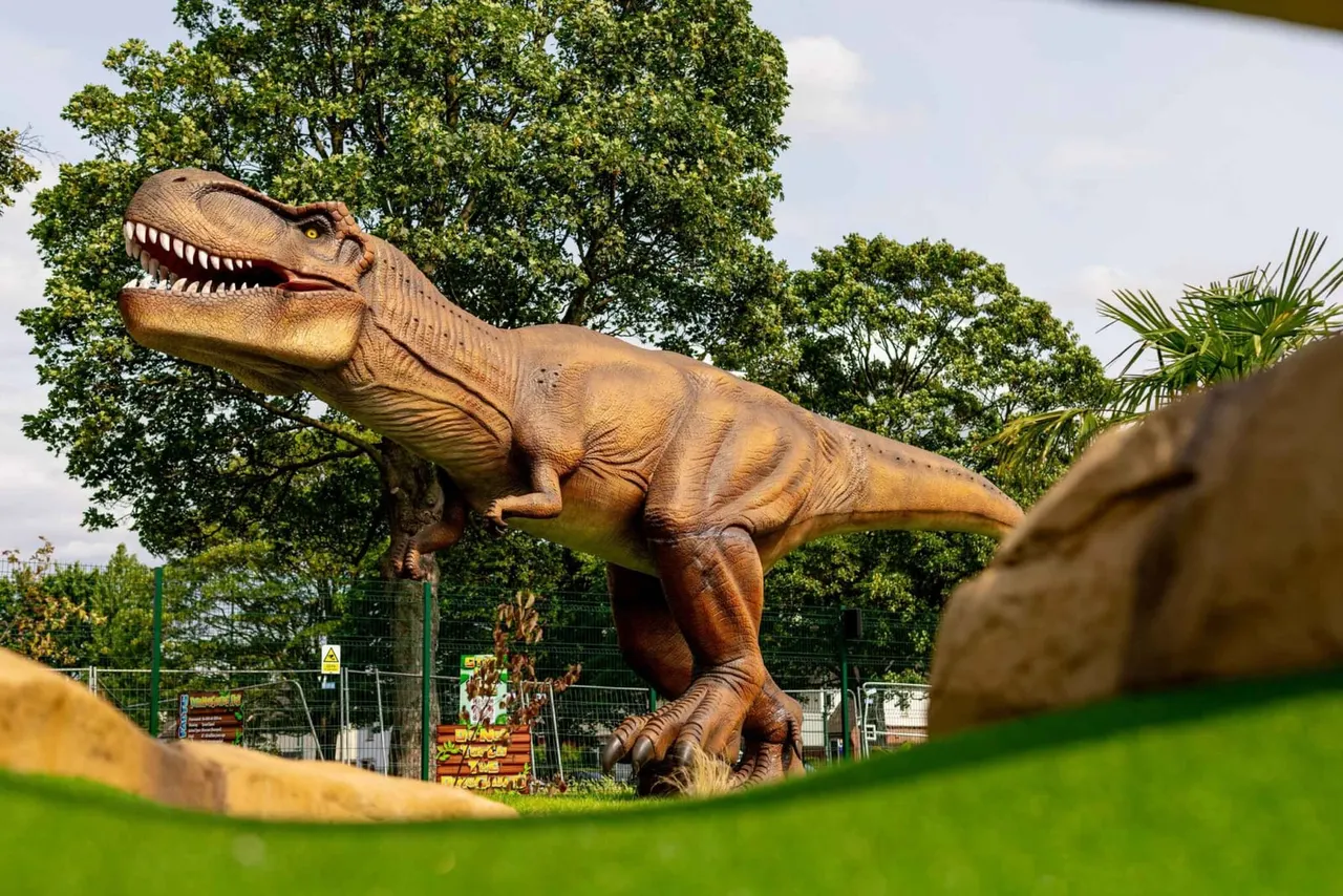 Large dinosaur statue with open mouth displaying teeth, surrounded by green lawn, trees and tropical plants in an outdoor adventure golf venue.