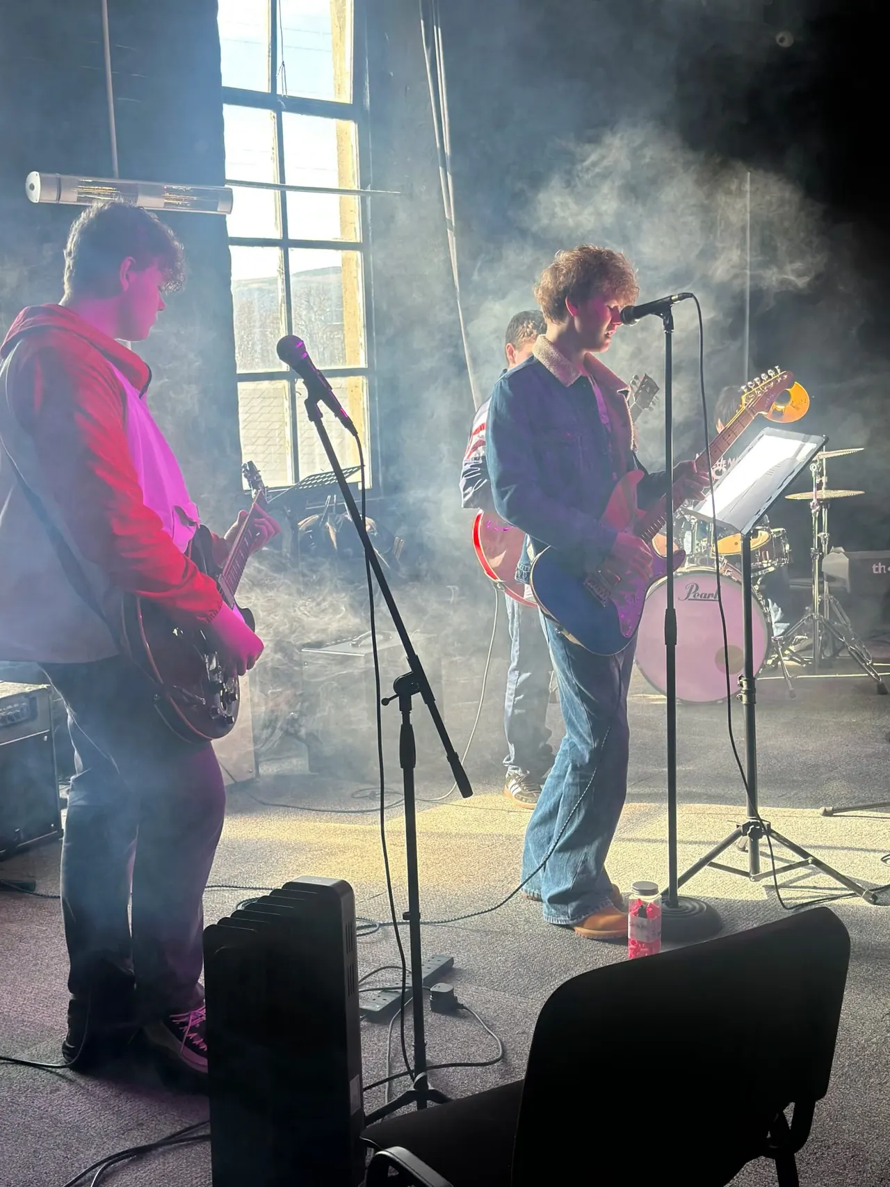 A band performs in a sunlit indoor space with stage smoke, featuring three musicians playing guitars and singing at microphones, with drums visible in the background.