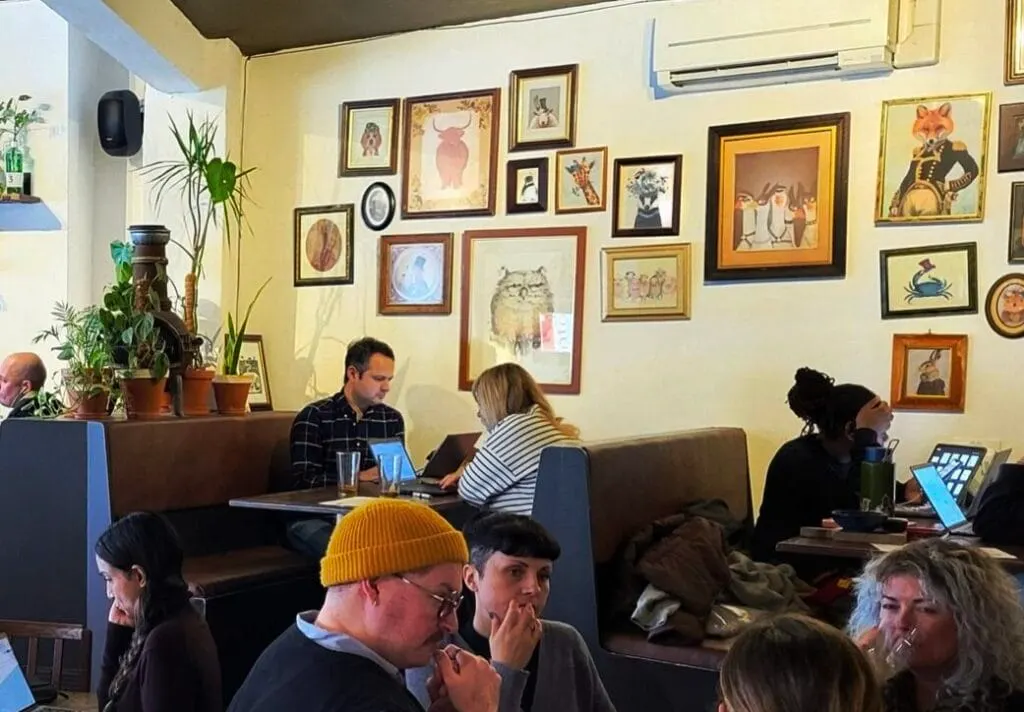 People seated on sofas in a café interior with yellow walls decorated with framed artworks and potted plants.