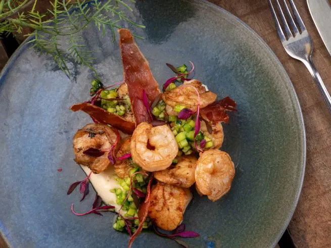 Cooked prawns and vegetables artfully plated on a blue-grey dish with fork and garnish visible.