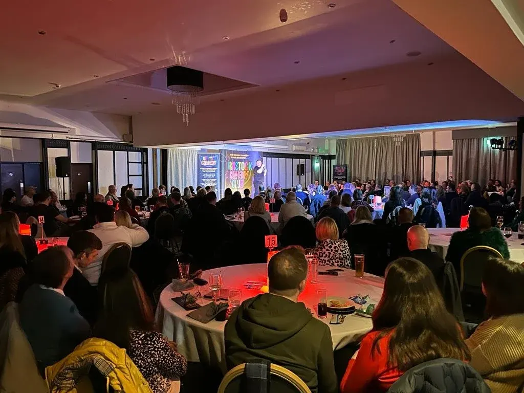 A large indoor event with hundreds of guests seated at round tables with red lighting, facing a stage with performers and screens.