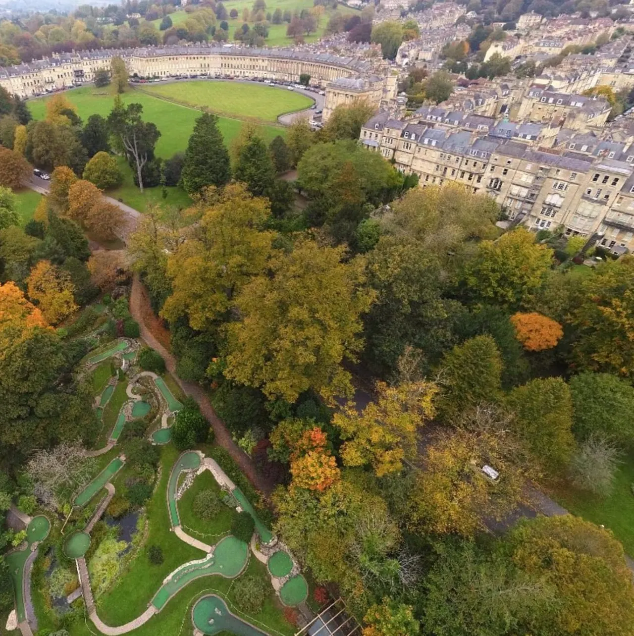 Aerial view of miniature golf course surrounded by autumn trees, with Georgian crescent buildings and parkland beyond.