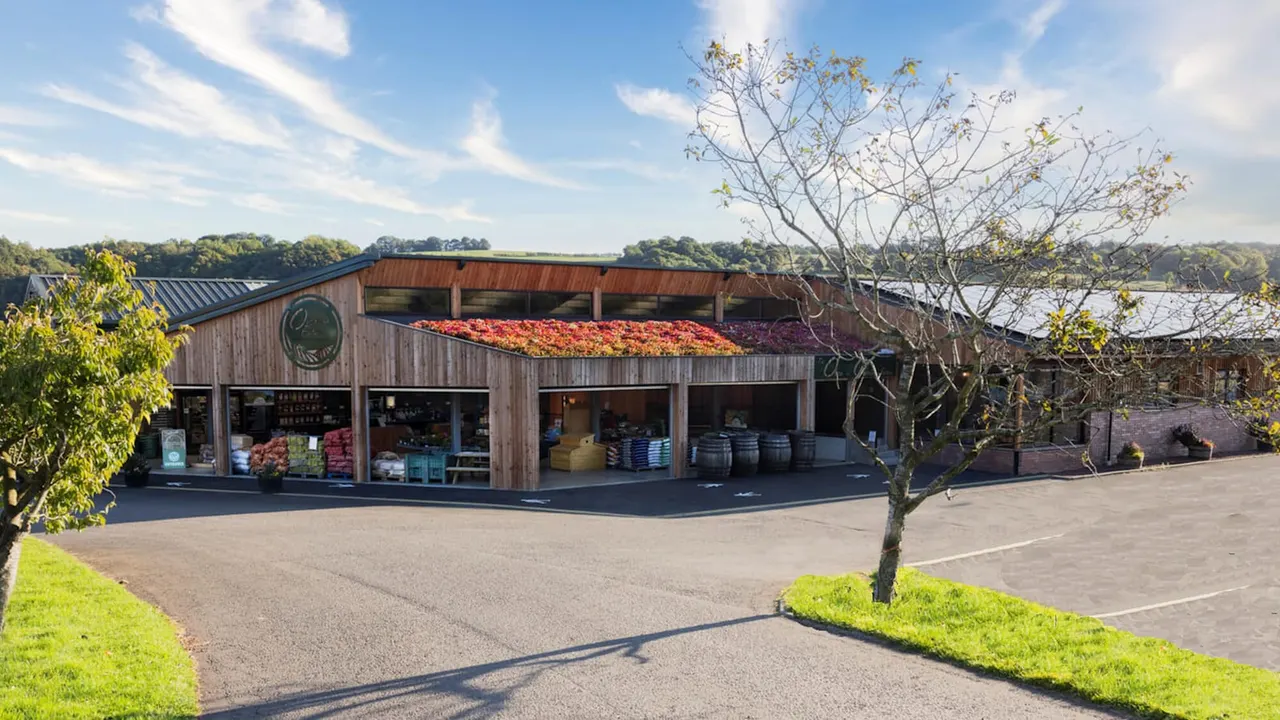 Wooden farm building with large windows displaying fresh produce, red apples drying on the roof, overlooking countryside and water.