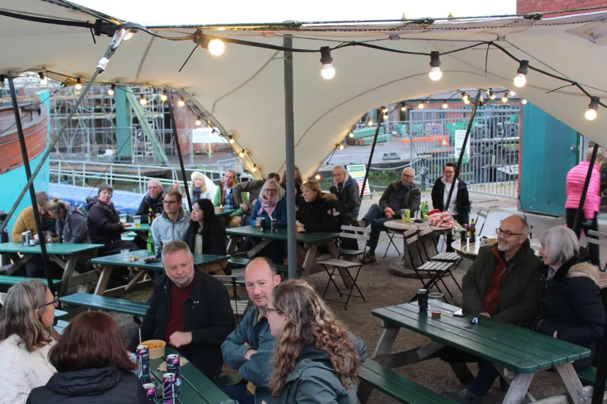 Groups of visitors seated and standing under a lit marquee at an outdoor waterside venue, with picnic tables and fairground rides visible beyond.