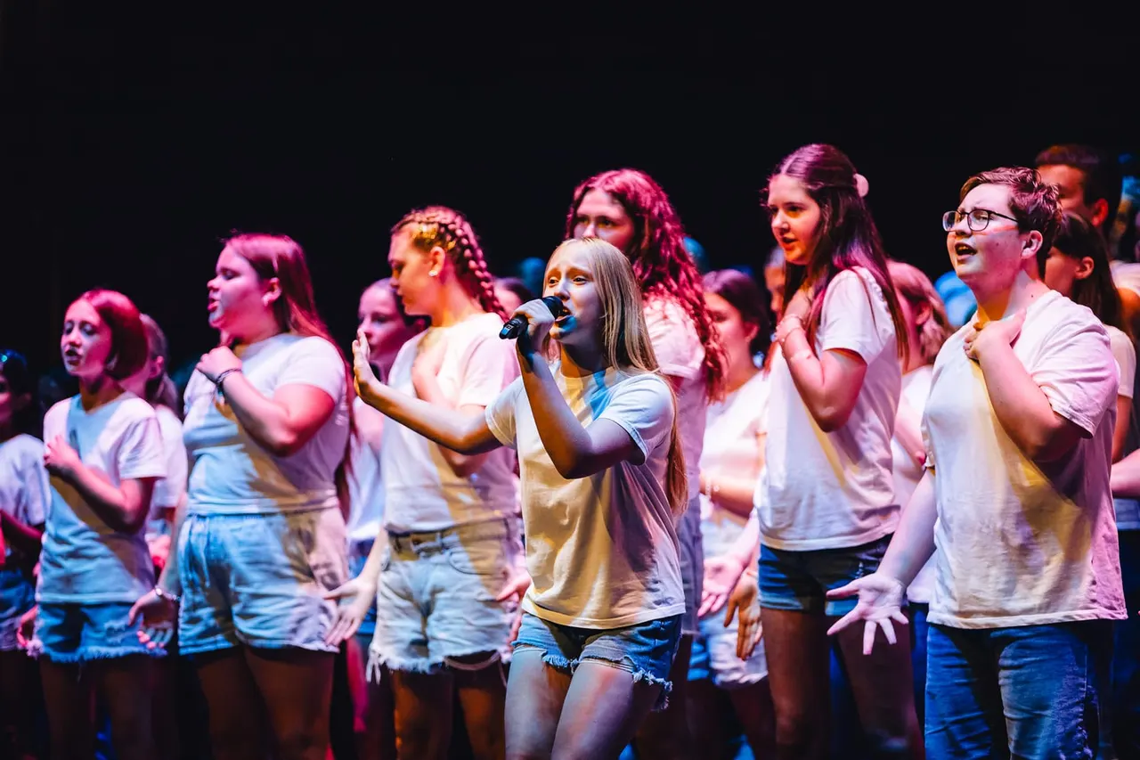 Group of young singers performing on stage with microphones under pink and blue stage lighting.