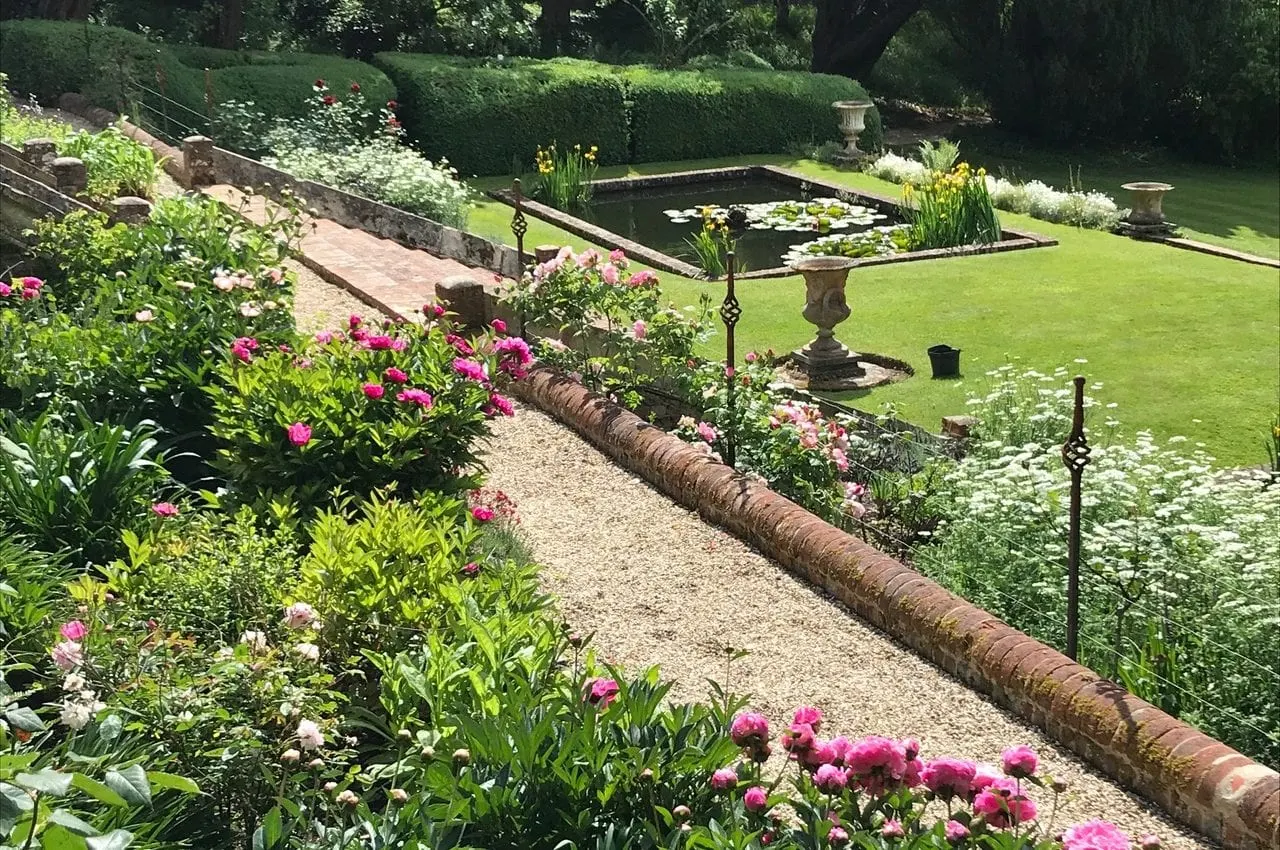 Formal gardens with pink flowering plants alongside a gravel path, leading to a rectangular pond surrounded by manicured hedges and lawn.