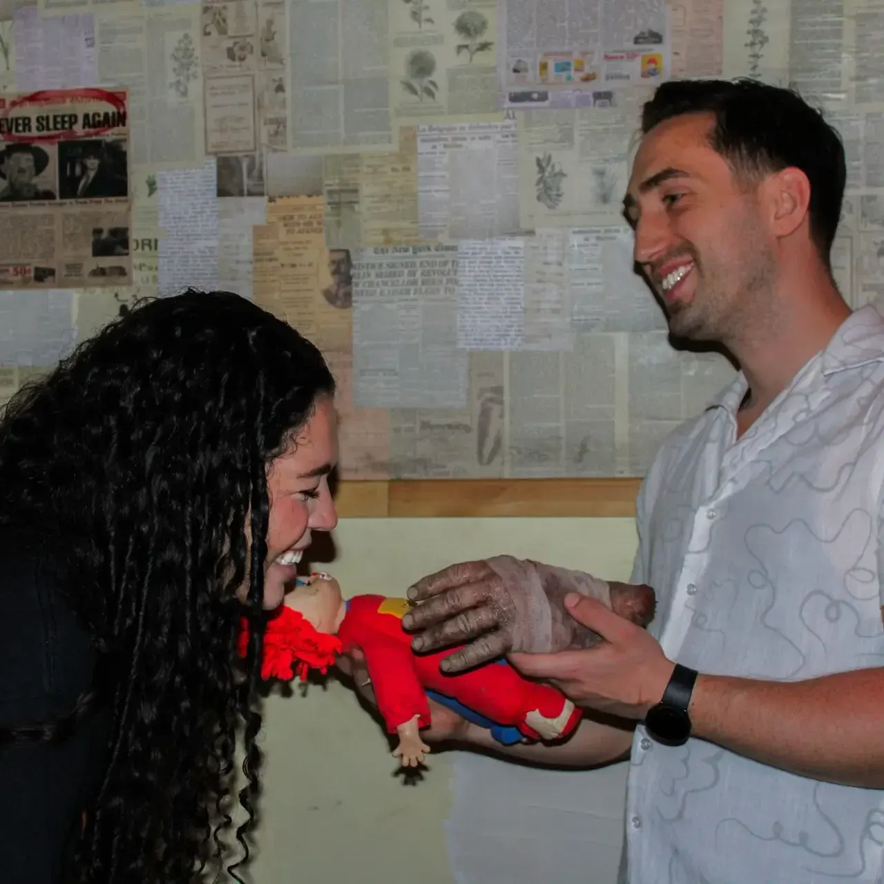 A man and woman smiling whilst holding a small doll in a room with newspaper clippings on the wall.
