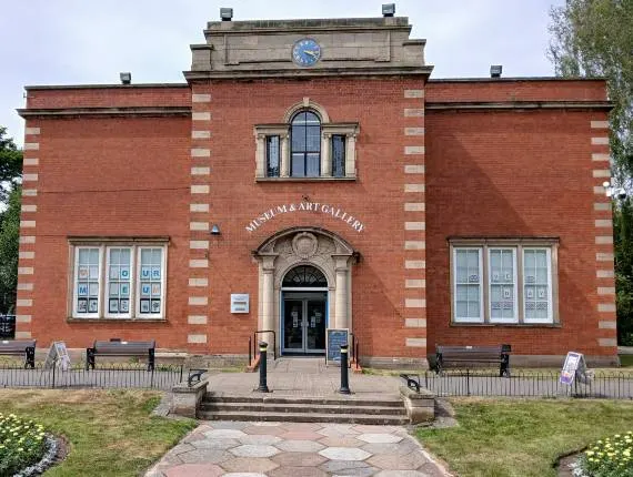 Exterior of a red brick museum building with a clock tower, arched entrance doorway, and manicured gardens.