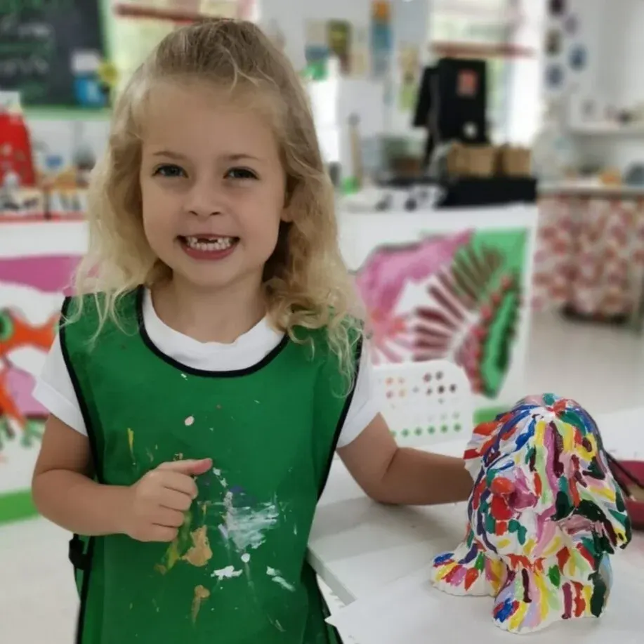 Young child in green apron smiling at camera whilst holding a colorful hand-painted ceramic piece in an art studio.