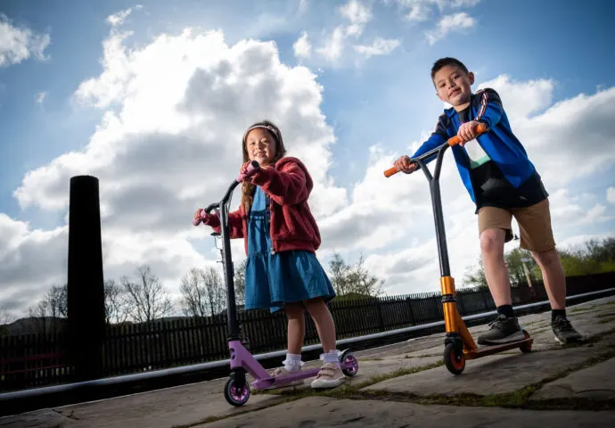 Two children riding scooters outdoors on a paved area beneath a blue sky with white clouds.